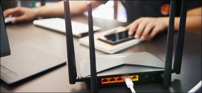 เราเตอร์ไร้สายบนโต๊ะกับผู้ชายที่ใช้คอมพิวเตอร์เดสก์ท็อป Mac และ iPhone ในพื้นหลัง