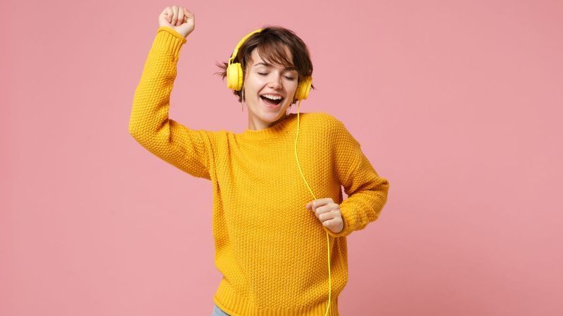 Frau im gelben Pullover tanzt beim Musikhören über gelbe, kabelgebundene Kopfhörer