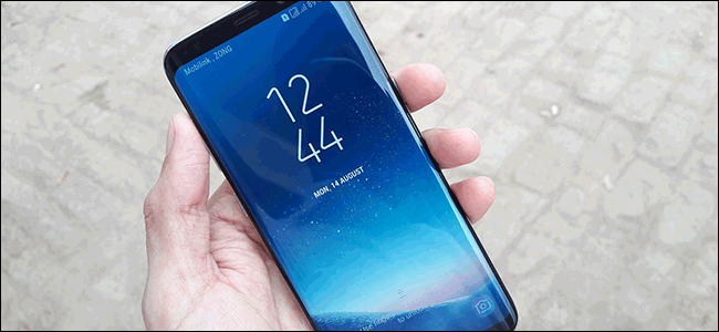 Roka, kas tur Samsung Galaxy S8 ar skārienekrānu, kurā ir redzams laiks un datums.