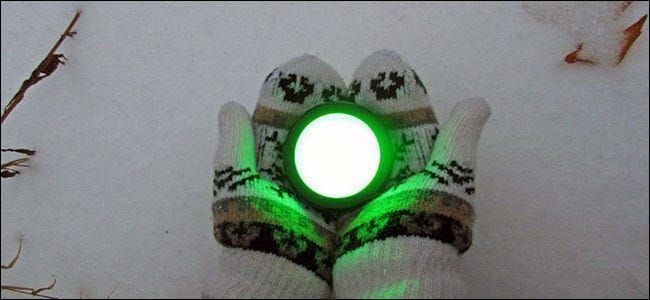 Un par de manos enguantadas sosteniendo un botón de eco que brilla intensamente verde sobre la nieve.