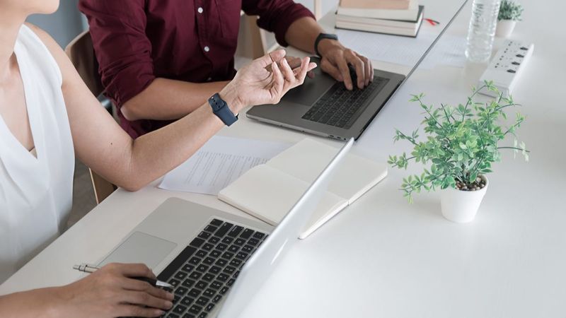 Zwei Personen mit zwei Laptops an einem Schreibtisch.