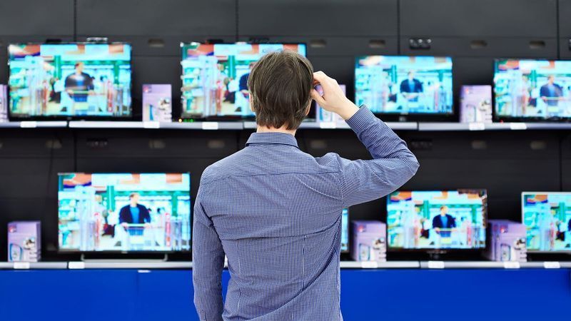 Un home que es rasca el cap mentre mira una pantalla de televisors en una botiga.